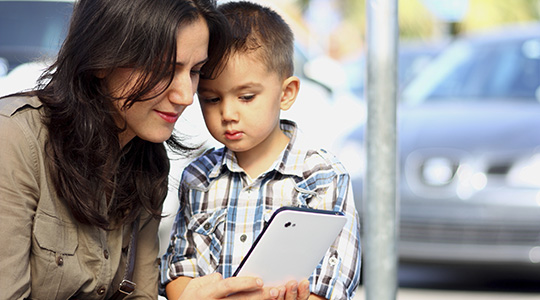 A young mother and son look at a smartphone screen together, with a parking lot visible through a window behind them.