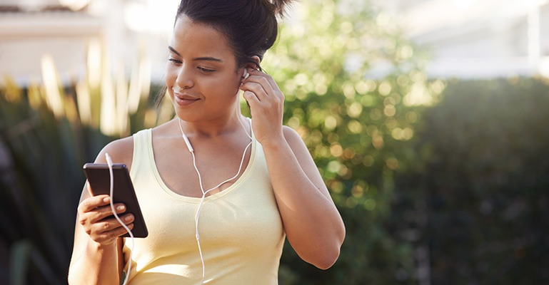 A young woman out for a walk looks at her phone screen and listens using headphones.