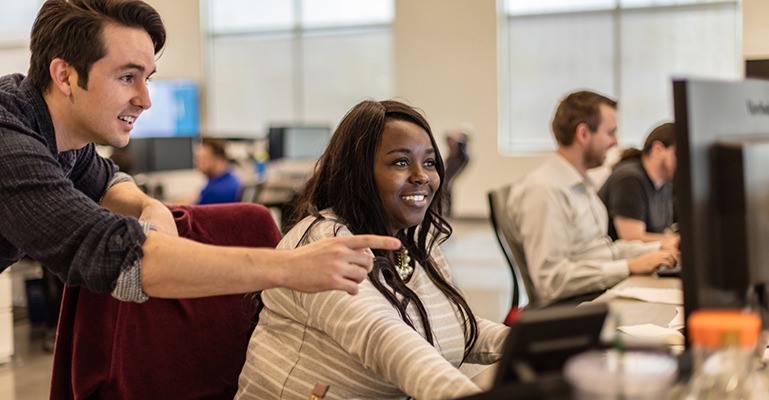 Two coworkers, one seated and one standing behind, look at a computer screen and smile.