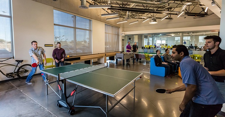 Four young men play ping-pong in the foreground with numerous others seated or standing in the background.