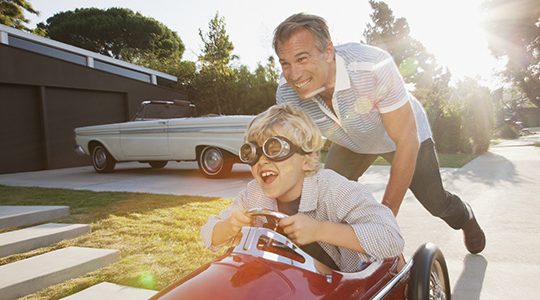 A smiling and laughing child rides in a toy car with an older man pushing it forward.