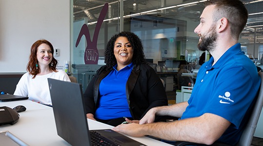 Three UnitedHealthcare Health Advocates smile and talk to each other behind a desk while waiting for a customer to call.