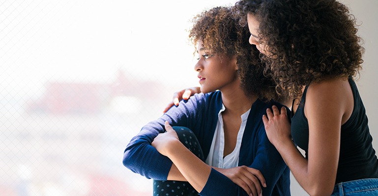 A young woman comforts a friend who looks out over the landscape, frowning.