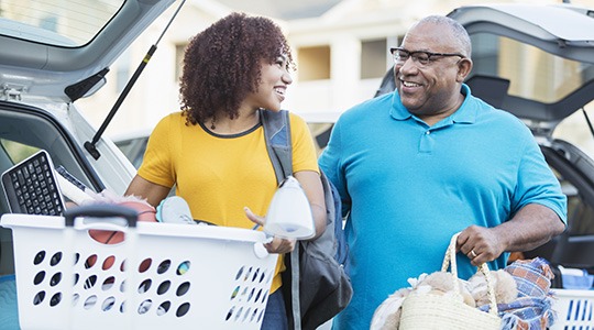 A smiling man and woman carry baskets of miscellaneous items out of the backs of two cars.