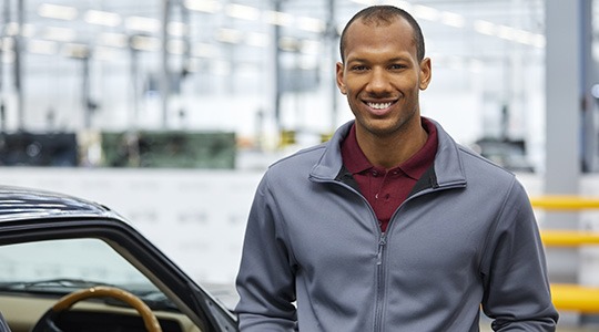 A smiling man in a zip-up jacket stands next to a car, with a cityscape in the background.