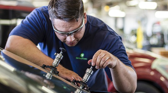 A man wearing goggles in a body shop works on removing a dent from a car.